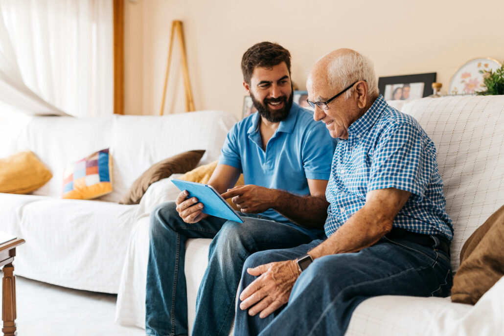 older man and caregiver sitting on couch with tablet